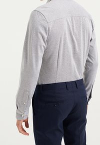 Man som bär en ljusgrå texturerad långärmad skjorta instoppad i marinblå dressbyxor, sedd från ryggen mot en neutral bakgrund.