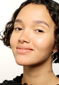 Close-up van een gezicht met krullend haar, een gouden septumring, glanzende lippen en een golvende zilveren oorbel. De huid lijkt glad met een natuurlijke glans.