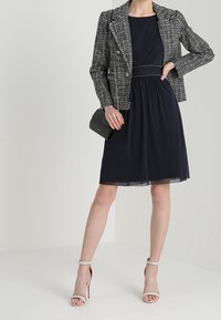 Navy Kleid mit taillierter Silhouette, kombiniert mit einem schwarz-weißen karierten Blazer. Dazu ein Clutch und High Heels mit Riemchen.
