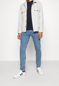 Ljusgrå skjortjacka med två bröstfickor, mörkblå t-shirt, blå skinny jeans och vita sneakers med svarta detaljer.