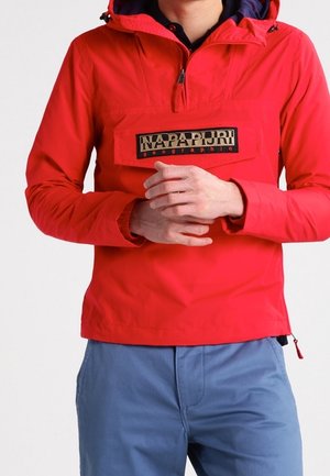 Personne portant une parka rouge Napapijri avec un logo sur la poche avant et un pantalon bleu, les mains jointes devant.