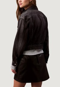 Černá džínová bunda s krátkým střihem, výraznými švy a manžetami na knoflíky, v kombinaci s žebrovaným šedým svetrem a černou koženou sukní.