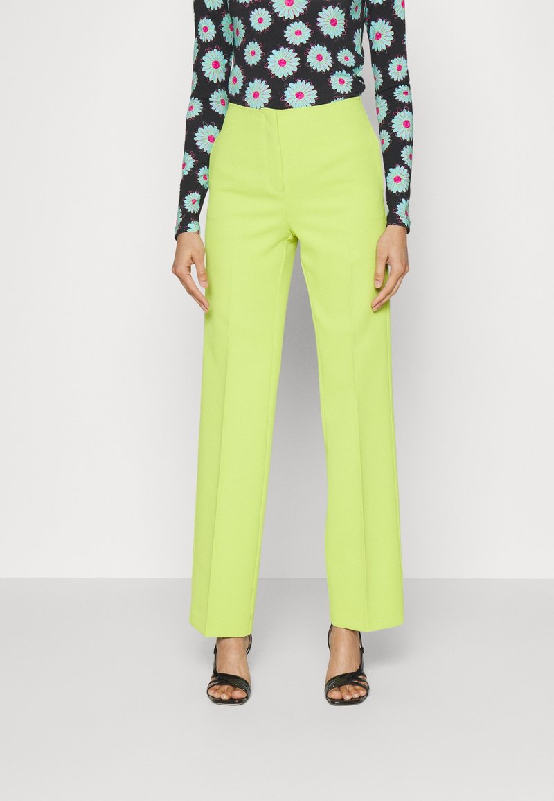 Femme portant un pantalon large vert citron, un haut noir à manches longues avec de grandes fleurs bleues et roses, et des sandales noires à bout ouvert.