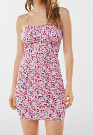 Vrouw in een mouwloze, roze, bloemen mini-jurk met dunne bandjes, staand tegen een effen witte achtergrond.