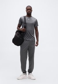 Άνδρας που φοράει γκρι αθλητική στολή και λευκά αθλητικά παπούτσια, κρατώντας μαύρη τσάντα duffel στον ώμο, στέκεται μπροστά σε λευκό φόντο.