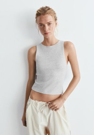 Jeune femme à la peau claire, avec des taches de rousseur et des cheveux blonds, portant un haut gris sans manches et un pantalon crème à cordon, debout contre un mur blanc.