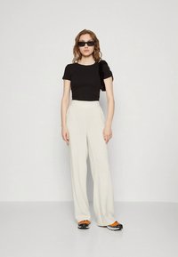 T-shirt preta cropped, calças largas cor de bege e ténis pretos com detalhes em laranja. Modelo usa óculos de sol e tem cabelo ondulado. Design simples.