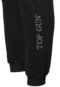 Schwarze Sweatpants aus weichem Stoff mit gerippten Bündchen, die an der Seite mit auffälliger silberner "TOP GUN" Stickerei verziert sind.