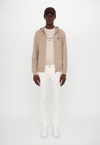 Sudadera beige con cremallera y cordones, camiseta blanca roto con texto, jeans blancos y zapatillas blancas. Gorra marrón completa el conjunto.