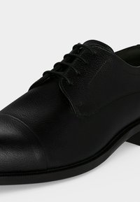 Chaussure habillée en cuir noir avec surface texturée, bout arrondi et lacets noirs sur fond blanc.