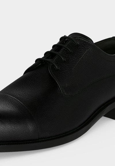 Chaussure habillée en cuir noir avec surface texturée, bout arrondi et lacets noirs sur fond blanc.