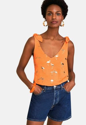 Naine kannab oranži värvi varrukateta t-särki flamingode trükiga ja seotud õlapaeladega, millele on lisatud tumedasinised teksapüksid ja rõngakujulised kõrvarõngad.