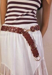 Cintura in pelle marrone con anelli circolari e hardware dorato, indossata sopra una gonna plissettata bianca con una finitura texture. Parte superiore a righe visibile sopra.