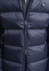 Mørkeblå dunjakke med horisontale quiltede seksjoner, doble glidelåser og logo-patch. Laget av lett, glatt stoff.