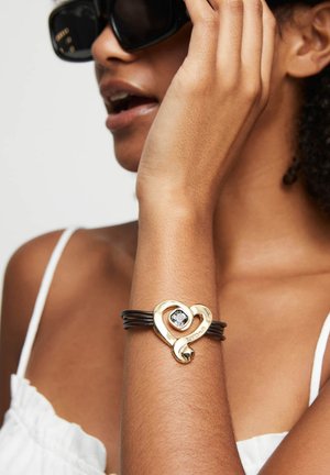 Pulsera de oro en forma de corazón con una piedra clara central, con correas de cuero oscuro. Superficie lisa con un acabado pulido y un diseño intrincado.