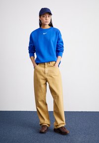 Blå sweatshirt, beiga lösa byxor, mörka sneakers och en marinblå keps. Sweatshirten har en Nike-logotyp och stora hängörhängen.