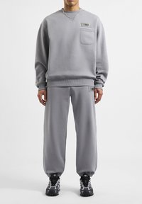 Grijze sweatshirt met een zak en logo, gecombineerd met bijpassende grijze joggingbroeken. Beide hebben een gladde textuur. Zwarte sportieve schoenen met een gemusterd ontwerp.