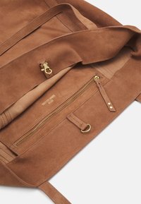 Brown suede tote bag interior with a zipper pocket, gold hardware, and a smooth texture. Accents include a strap and branding inside.