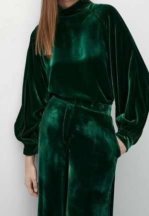 Femme portant une tenue en velours vert émeraude avec de longues manches bouffantes et un pantalon taille haute, debout devant un fond blanc uni.