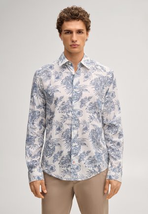 Mężczyzna w białej koszuli z długim rękawem z niebieskim kwiatowym wzorem i beżowych spodniach, stojący na jednolitym tle.