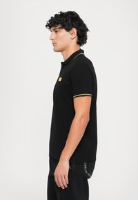 Camisa polo preta com detalhes amarelos na gola e nas mangas, apresentando um pequeno logo na parte da frente. Confeccionada em tecido de algodão suave.