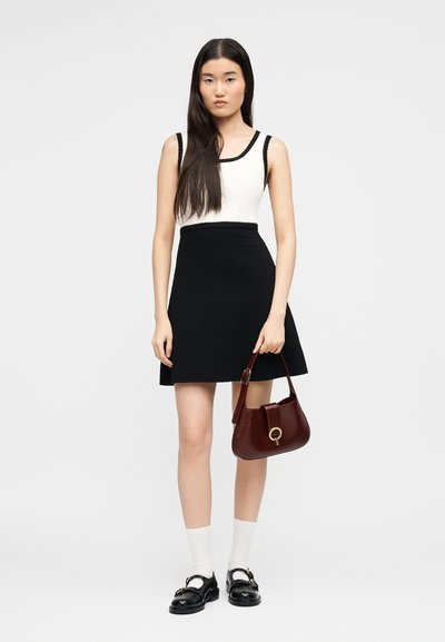 Νεαρή γυναίκα με μακριά μαύρα μαλλιά, φορώντας λευκό τοπ, μαύρη φούστα, άσπρες κάλτσες, μαύρα παπούτσια, κρατώντας καφέ τσάντα, σε απλό φόντο.