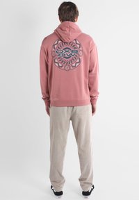 Felpa color rosa con cappuccio, caratterizzata da una grafica circolare e testo sulla schiena, abbinata a pantaloni in corduroy beige e scarpe nere.