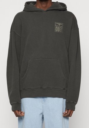 Tumman oliivinvihreä huppari, jossa edessä tasku ja vasemmassa rinnassa pieni "OBEY"-logo, yhdistetty vaaleansinisiin farkkuihin.