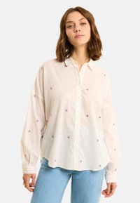 Camicia bianca a maniche lunghe con colletto a bottoni, decorata con piccoli fiori ricamati in rosso e blu. Tessuto morbido e leggero con una vestibilità comoda.