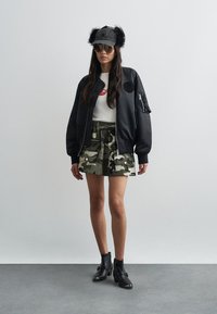 Jachetă bomber neagră cu un patch rotund, tricou alb cu grafică, fustă camuflaj și ghete negre până la gleznă. Accesorii includ ochelari de soare și o căciulă neagră din blană.