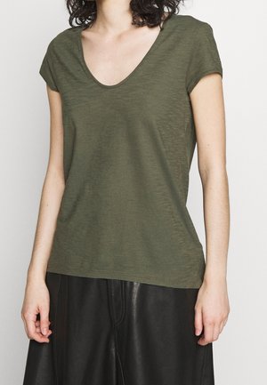 Kobieta ubrana w oliwkowozielony T-shirt z krótkim rękawem i dekoltem w serek oraz czarną skórzaną spódnicę, stojąca na tle jednolitego białego tła.