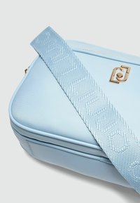 Svijetloplava torba preko ramena s glatkom teksturom, zlatnim detaljima i remenom s uzorkom koji sadrži ime branda. Zipper zatvaranje na vrhu.