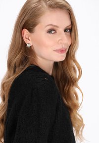 Sort, struktureret sweater med rund hals; modellen har langt, bølget hår og bærer firkantede, sølvfarvede stud ørestikker med en poleret finish.