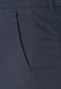 Tmavě modré látkové kalhoty s bočními kapsami, slim fit a hladkou texturou s viditelnými švy.
