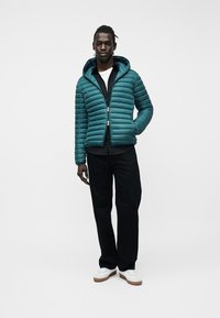 Άνδρας που φοράει πετρόλ καπιτονέ μπουφάν με κουκούλα, μαύρο παντελόνι, μαύρο πουκάμισο, λευκό μπλουζάκι από μέσα και λευκά αθλητικά παπούτσια με καφέ σόλες, όρθιος.