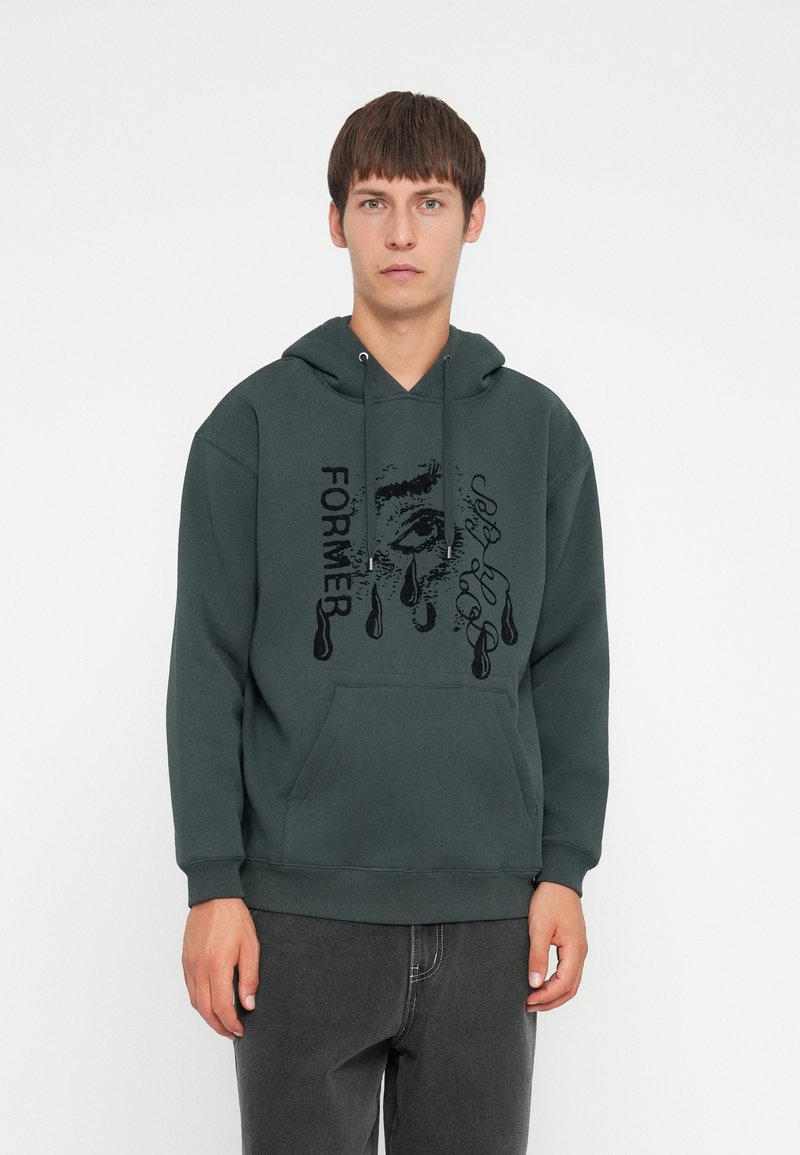 Young man wearing dark green hoodie with black graphic of an eye and the words "FORMER" and "Stay Lost" on front.