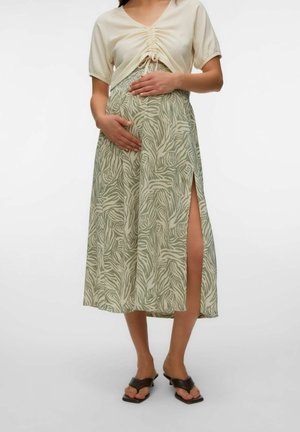 Personne enceinte portant un haut plissé clair et une jupe à motif feuilles avec une fente frontale sur la cuisse, tenant son ventre avec les deux mains.