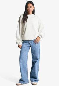 Una donna si erge con sicurezza indossando una felpa bianca e jeans a gamba larga di un blu chiaro, con lunghi capelli scuri e in una posa rilassata.