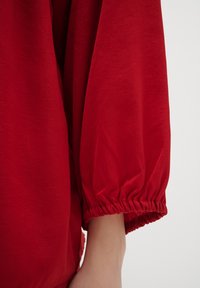 Czerwony, lekki top z tkaniny o luźnym kroju i objętościowych rękawach z elastycznymi mankietami, charakteryzujący się gładką teksturą i subtelnym połyskiem.