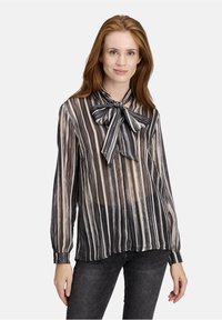 Vrouw met lang rood haar, gekleed in een zwart-wit-bruin verticaal gestreepte blouse met een grote strik kraag en donkere jeans.