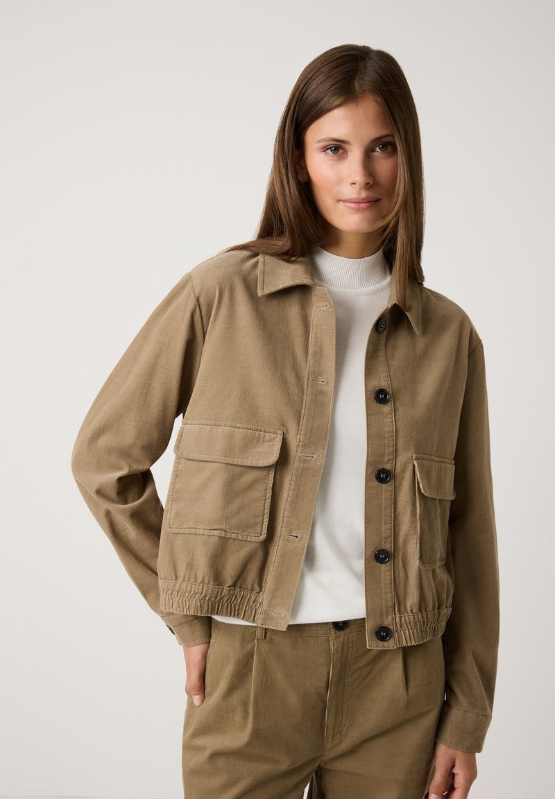 Khaki Baumwolljacke mit zwei vorderen Taschen, Knopfverschluss und elastischem Saum. Über einem weißen Oberteil getragen, was einen lässigen Schnitt zeigt.