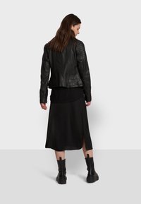 Veste de moto en cuir noir avec épaules matelassées, associée à une longue robe noire avec une fente sur le côté. Des bottes de combat noires épaisses complètent le look.