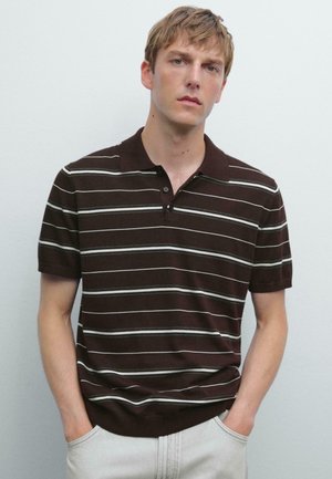 Jonge man met zijn handen in zijn zakken, gekleed in een donkerbruine polo met witte horizontale strepen tegen een effen lichte achtergrond.