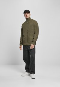 Oliven grønn strikket zip-up genser med høy krage og to frontlommer, kombinert med mørke jeans og hvite sneakers.