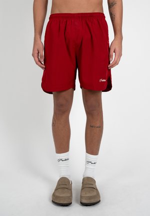 Uomo che indossa pantaloncini rossi con vita elastica, calzini bianchi a metà polpaccio con scritte nere e scarpe beige slip-on, in piedi su uno sfondo uniforme.