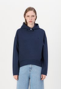 Felpa a cappuccio corta blu navy con chiusura a bottone, tessuto morbido e un logo sottile sul petto. Abbinata a jeans larghi azzurri.