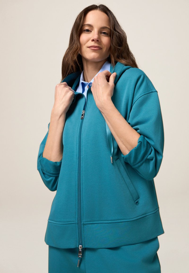 Sudadera con cremallera en color verde azulado, con una capucha espaciosa, mangas largas y un corte relajado. Cuenta con una cremallera frontal y cordones ajustables para mayor comodidad.