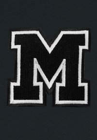 Lettera "M" nera ricamata con contorno bianco, su uno sfondo scuro. La texture è in rilievo, enfatizzando i dettagli del ricamo.