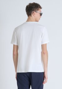 Camiseta blanca de algodón con mangas cortas, cuello redondo y un mínimo de marca en la parte posterior. Combinada con pantalones azul marino. Textura suave.