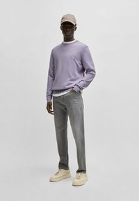 Helllila langärmliges Sweatshirt mit kleinem Logo, kombiniert mit grauen Jeans und cremefarbenen Sneakers sowie einer beigen Kappe, was einen lässigen Look vermittelt.